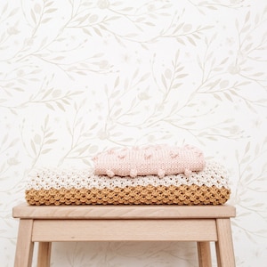 May include: A wooden stool with a light brown finish, topped with a white and beige crocheted blanket and a pink knitted blanket. The stool is in front of a white wall with a delicate pattern of beige branches and leaves.