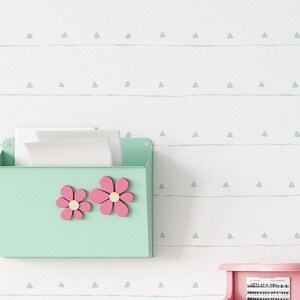 May include: A white wall with a light green and white geometric pattern wallpaper. A light green file holder with pink flowers is mounted on the wall. A pink toy piano sits on a white table with a magazine on top. Two star-shaped decorations are on the table.
