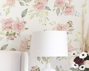 May include: A white bedroom with a white headboard, a white lamp with a white shade, and a white bedside table with a vase of flowers. The wall is covered in floral wallpaper featuring pink roses and green leaves.