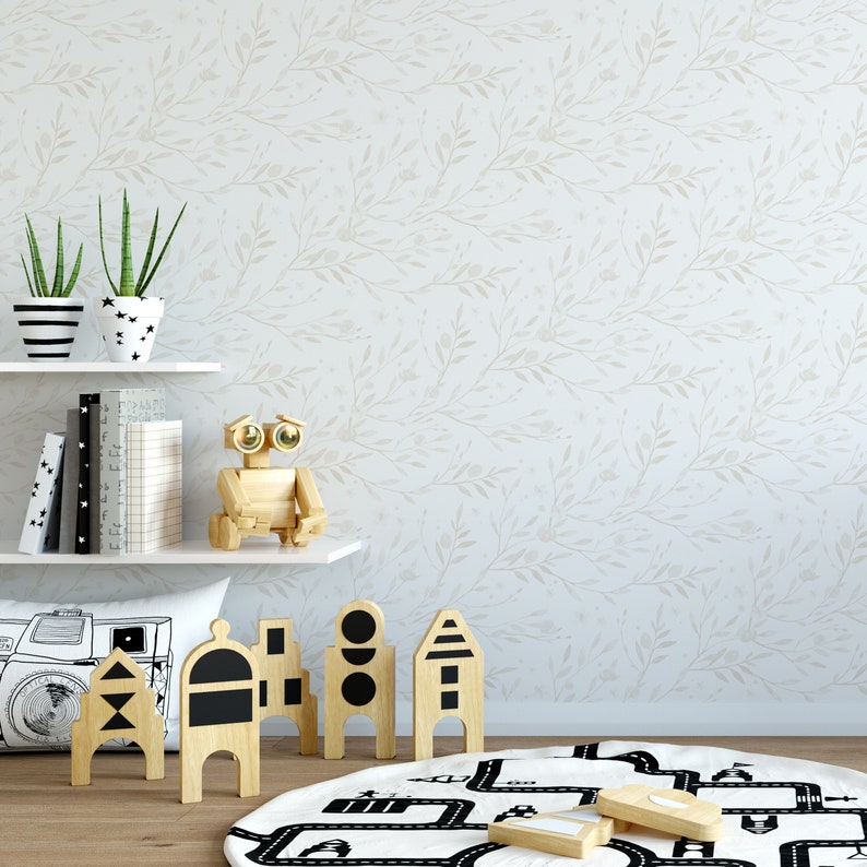 May include: A white wall with a light brown floral pattern. A wooden shelf with two potted plants, books, and a wooden robot toy. A white pillow with a camera graphic. A black and white play mat with a road design. Wooden toy blocks with black geometric shapes.