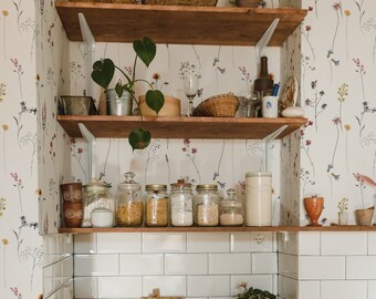 May include: A set of wooden shelves with white brackets, filled with various items, including jars of dried goods, a potted plant, and a wicker basket. The shelves are mounted against a white wall with a floral wallpaper pattern.
