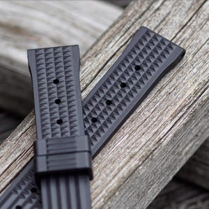 May include: Two black watch straps with a geometric diamond pattern are displayed on a weathered wooden surface. The straps have multiple holes for adjustment and a secure clasp. The textured design adds a modern touch to the watch accessory.