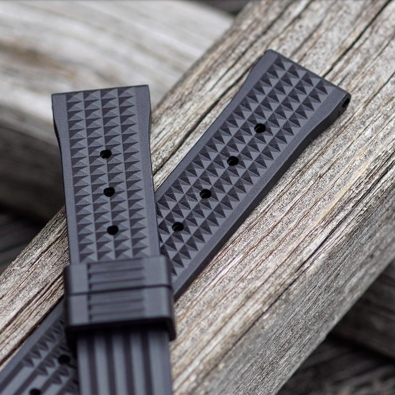 May include: Two black watch straps with a geometric diamond pattern are displayed on a weathered wooden surface. The straps have multiple holes for adjustment and a secure clasp. The textured design adds a modern touch to the watch accessory.