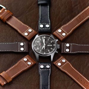May include: A silver chronograph watch with a black face and white markings. The watch is surrounded by eight different leather watch straps in various shades of brown and black.