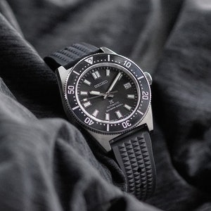 May include: A black Seiko automatic diver's watch with a black dial, a rotating bezel, and a black rubber strap. The watch face displays the date and luminous markers. The watch is set against a dark gray background.