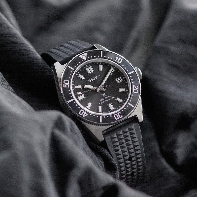 May include: A black Seiko automatic diver's watch with a black dial, a rotating bezel, and a black rubber strap. The watch face displays the date and luminous markers. The watch is set against a dark gray background.