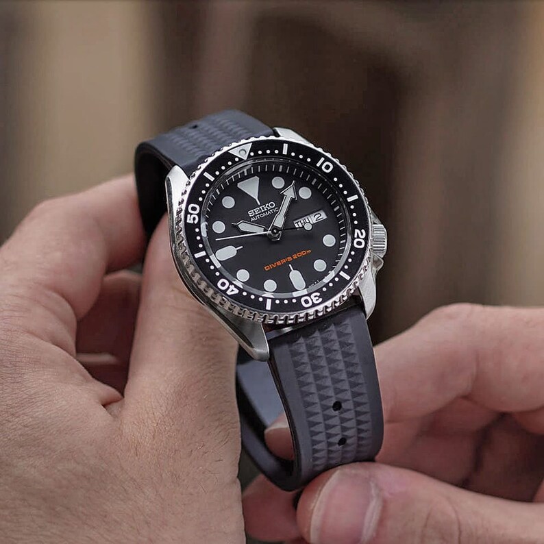 May include: A black-faced Seiko automatic dive watch with a silver bezel and a gray rubber strap. The watch features white hour markers, a day-date display, and the text "DIVER'S 200m" in orange. The strap has a geometric pattern.
