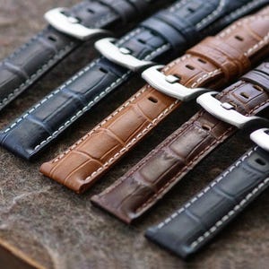 May include: Close-up of several leather watch straps in various colors, including black, navy, brown, and gray. Each strap features a crocodile pattern, white stitching, and a silver-colored buckle. The straps are arranged diagonally.