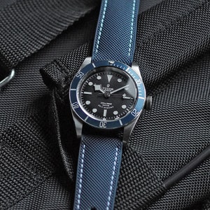 Puede incluir: Un reloj Tudor negro y plateado con un bisel azul y una correa de nailon azul y blanca a rayas. La esfera del reloj tiene marcas blancas y el texto "TUDOR" y "2000m-160ft" es visible.