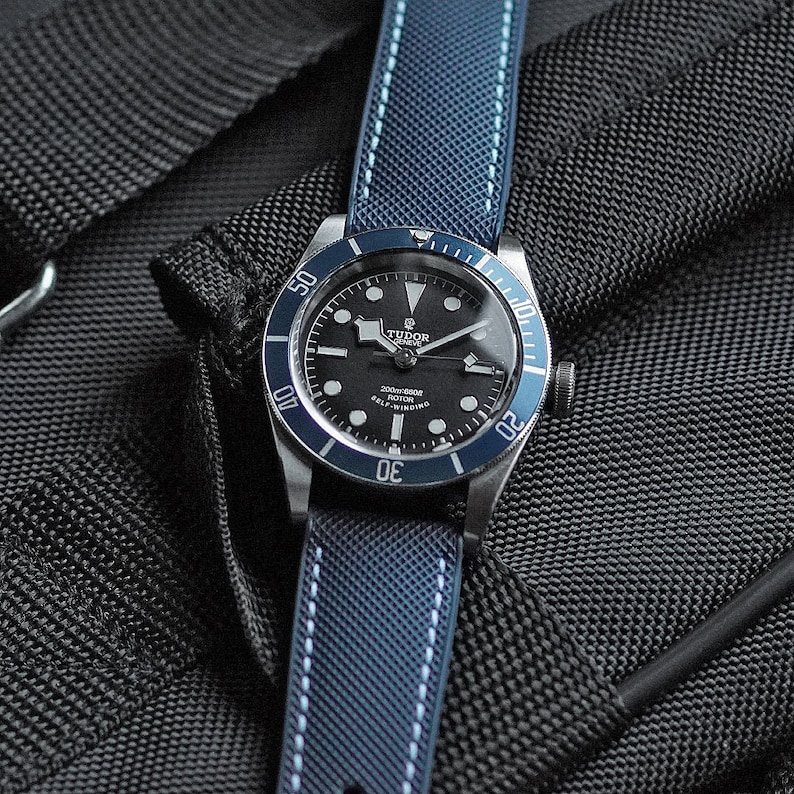 Puede incluir: Un reloj Tudor negro y plateado con un bisel azul y una correa de nailon azul y blanca a rayas. La esfera del reloj tiene marcas blancas y el texto "TUDOR" y "2000m-160ft" es visible.