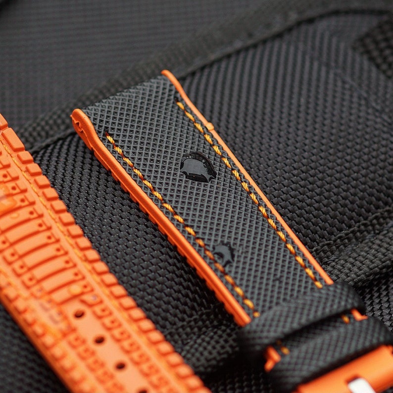 Puede incluir: Una correa de reloj negra con costuras naranjas. La correa tiene una superficie texturizada y est&aacute; hecha de un material duradero.