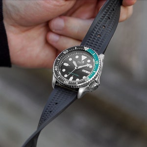 May include: A stainless steel wristwatch with a black dial, a green and black bezel, and a black rubber strap. The watch face displays the day and date. The strap is textured with a repeating pattern.