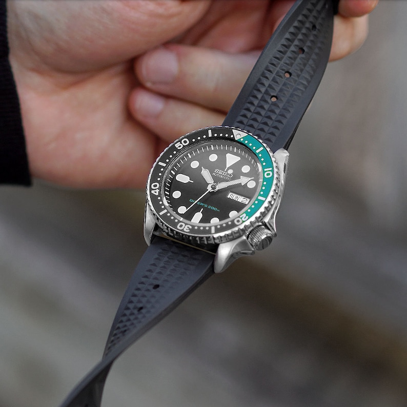 May include: A stainless steel wristwatch with a black dial, a green and black bezel, and a black rubber strap. The watch face displays the day and date. The strap is textured with a repeating pattern.