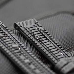 Puede incluir: Primer plano de una correa de reloj de caucho negro con una superficie texturizada. La correa tiene una serie de formas rectangulares en relieve.