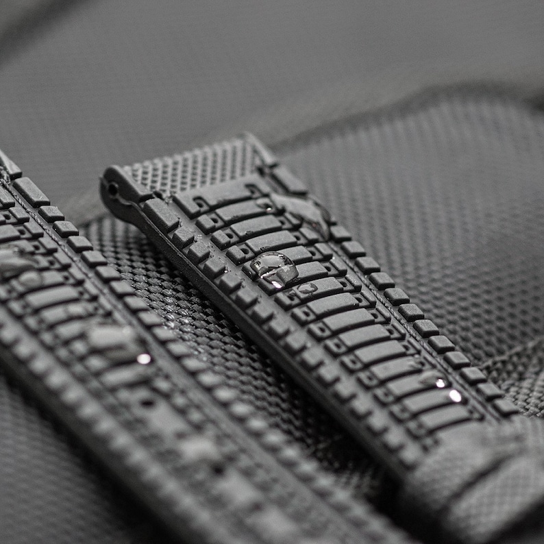 Puede incluir: Primer plano de una correa de reloj de caucho negro con una superficie texturizada. La correa tiene una serie de formas rectangulares en relieve.