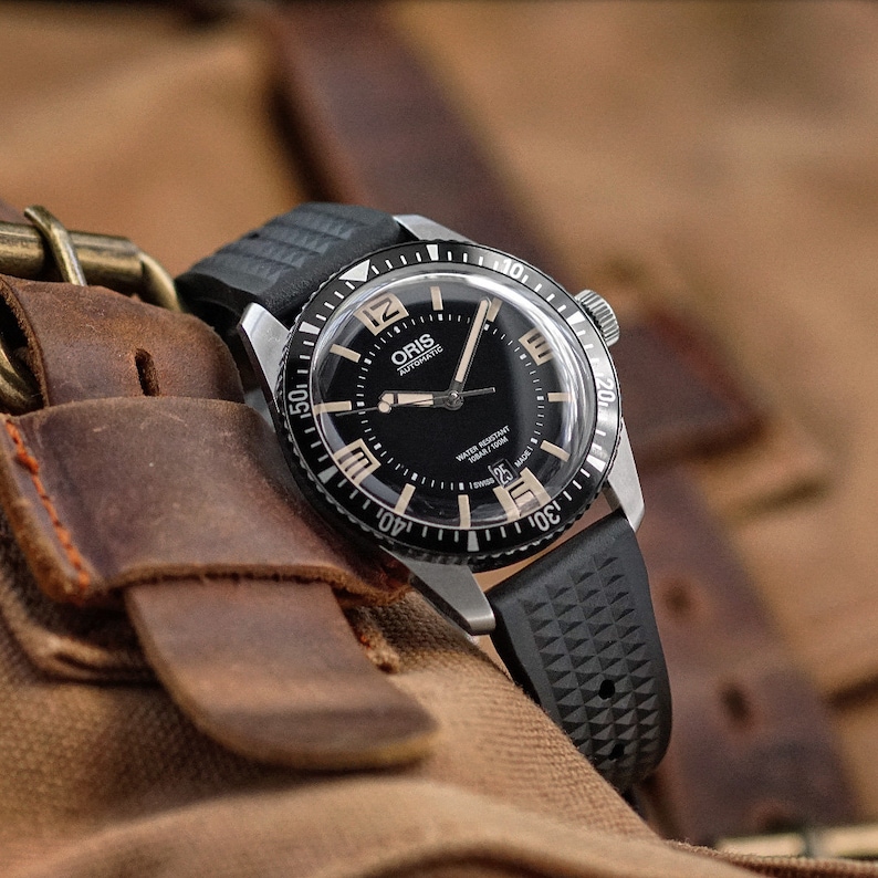 May include: A black and silver Oris automatic wristwatch with a black dial and a black rubber strap. The watch has a rotating bezel with beige numerals and is water-resistant to 100m. The watch is displayed on a brown leather surface.