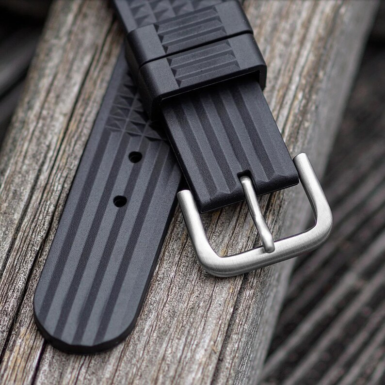 May include: Close-up of a black watch strap with a silver buckle. The strap has a textured, geometric design and vertical lines. The buckle is a brushed silver color. The strap is lying on a weathered wooden surface.