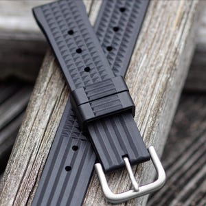 May include: Black watch band with a stainless steel buckle. The band is made of a textured material with a geometric pattern and vertical lines. The band is shown on a wooden surface.