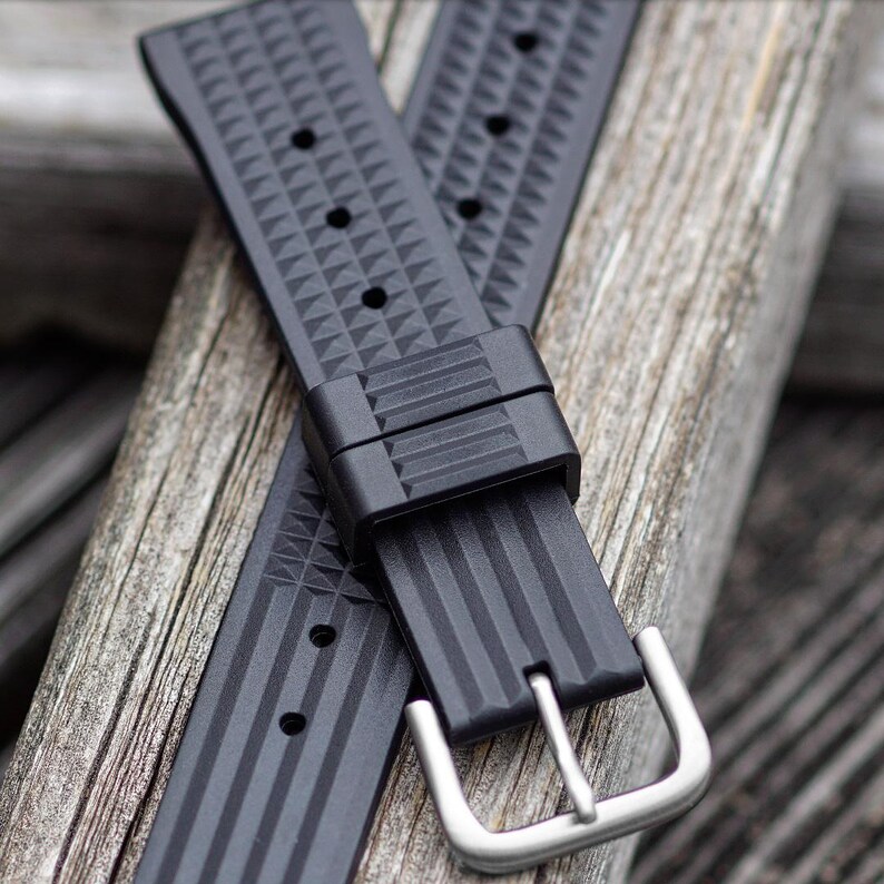 May include: Black watch band with a stainless steel buckle. The band is made of a textured material with a geometric pattern and vertical lines. The band is shown on a wooden surface.
