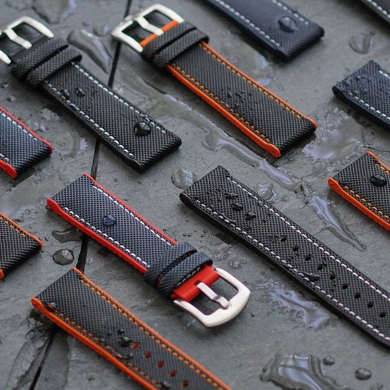 Puede incluir: Una colecci&oacute;n de correas de reloj negras con costuras naranjas. Las correas tienen una superficie texturizada y una hebilla plateada.