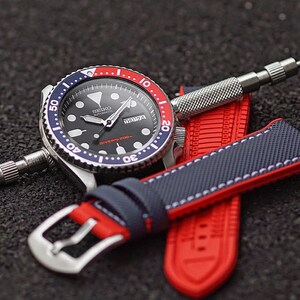 Puede incluir: Un reloj de buceo autom&aacute;tico Seiko negro con un bisel rojo y azul. El reloj est&aacute; en una correa de silicona roja y azul con costuras blancas. La esfera del reloj tiene una ventana de fecha y una ventana del d&iacute;a de la semana. El reloj est&aacute; sobre una superficie negra.
