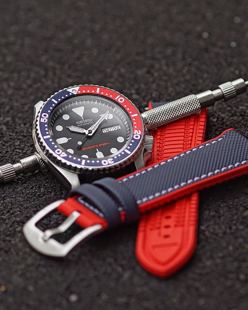 Puede incluir: Un reloj de buceo autom&aacute;tico Seiko negro con un bisel rojo y azul. El reloj est&aacute; en una correa de silicona roja y azul con costuras blancas. La esfera del reloj tiene una ventana de fecha y una ventana del d&iacute;a de la semana. El reloj est&aacute; sobre una superficie negra.