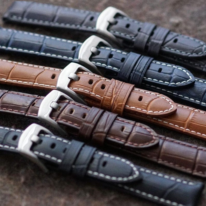 May include: Close-up of several leather watch straps in various colors, including brown, navy, and gray. Each strap features a textured, crocodile-like pattern, white stitching, and a silver-colored buckle. The straps are arranged in a diagonal row.