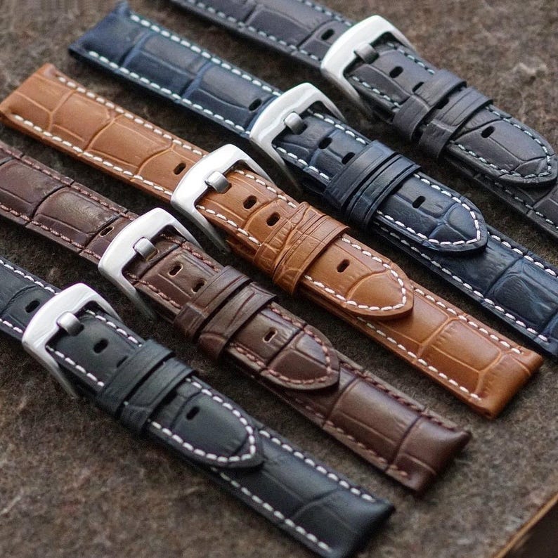 May include: A close-up shot of several watch straps in various colors, including navy blue, brown, and black. The straps have a textured, crocodile-like pattern and white stitching. Each strap features a silver buckle.