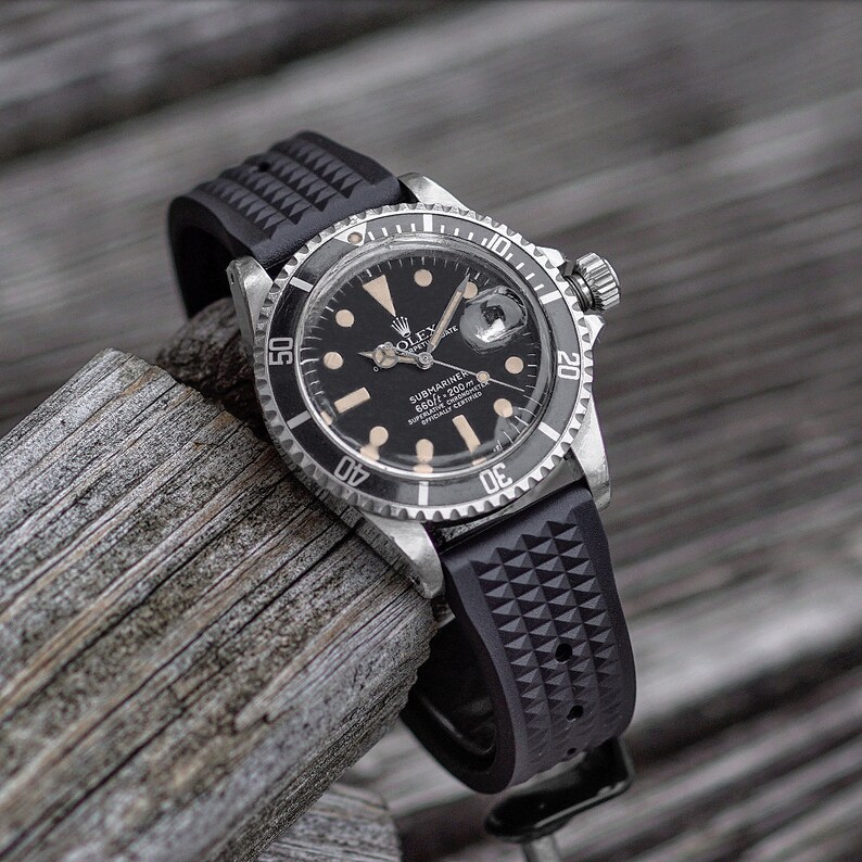 May include: A black Rolex Submariner watch with a black rubber strap. The watch has a black dial with luminous markers and hands. The bezel is silver with black markings. The watch is displayed on a piece of weathered wood.