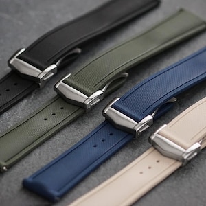 May include: Four watch straps in black, olive green, navy blue, and beige. Each strap features a textured surface, a series of adjustment holes, and a polished silver-tone metal deployment clasp mechanism for securing the watch to the wrist.