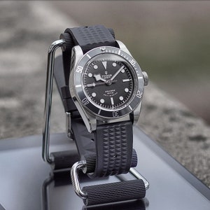 May include: A black and silver wristwatch with a black dial and a black rubber strap. The watch has a rotating bezel and is displayed on a silver stand. The watch face has white hour markers and hands.