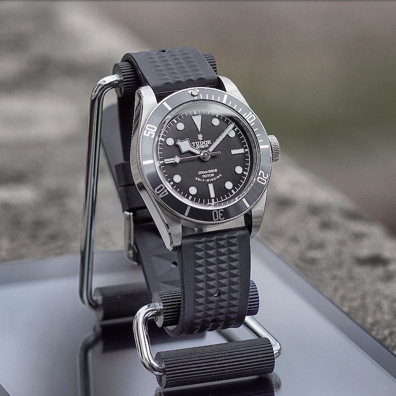 May include: A black and silver wristwatch with a black dial and a black rubber strap. The watch has a rotating bezel and is displayed on a silver stand. The watch face has white hour markers and hands.