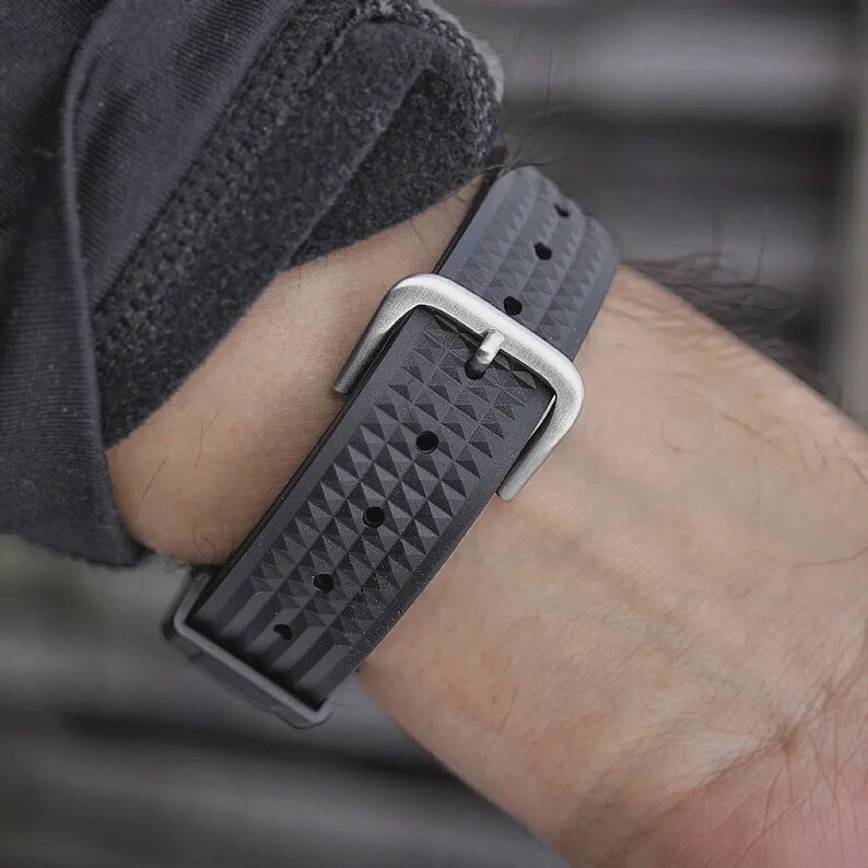 May include: Close-up of a black watch strap with a geometric pattern and a silver buckle. The strap is secured on a wrist, showcasing the textured design and the metallic clasp.