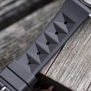 May include: Close-up of a black watch strap with a geometric pyramid design. The strap is made of a flexible material and features several holes for adjustment. The watch strap is resting on a weathered wooden surface.