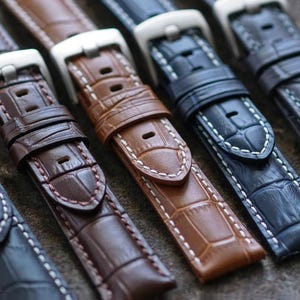 May include: Close-up of several leather watch straps in various shades of brown and blue. The straps feature a crocodile pattern, white stitching, and silver buckles. The straps are arranged side by side.