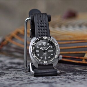 May include: A black and silver Seiko PADI automatic diver's watch with a black rubber strap. The watch face is black with white markings and hands. The watch is displayed on a silver stand.