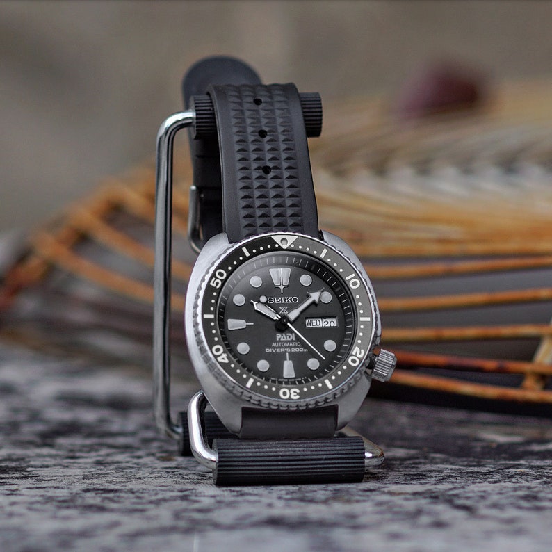 May include: A black and silver Seiko PADI automatic diver's watch with a black rubber strap. The watch face is black with white markings and hands. The watch is displayed on a silver stand.