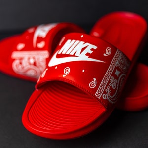 May include: Red Nike slides with a white paisley pattern and the Nike logo. The slides have a textured footbed and are set against a dark background. The slides are a vibrant red color.
