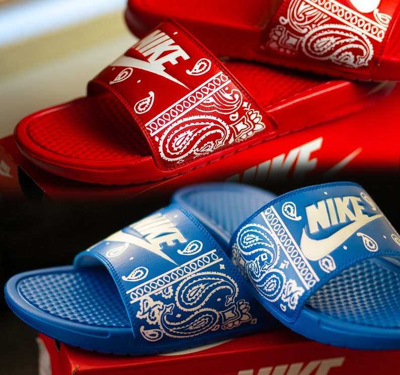 May include: Red and blue Nike slides with white paisley bandana print straps. The Nike logo is visible on the straps. The slides are displayed on top of red Nike boxes. The slides are a casual footwear item.
