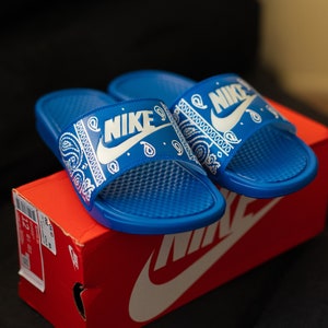 May include: A pair of blue Nike slides with a white paisley design and the Nike logo. The slides are resting on a red Nike shoe box. The slides have a textured footbed.