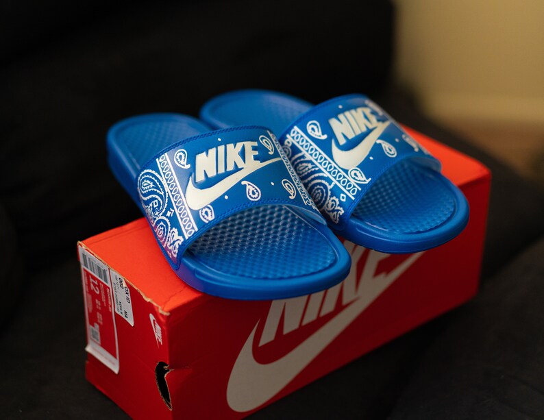 May include: A pair of blue Nike slides with a white paisley design and the Nike logo. The slides are resting on a red Nike shoe box. The slides have a textured footbed.