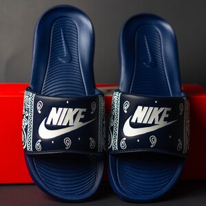 May include: A pair of navy blue Nike slides. The slides feature a white paisley pattern on the strap with the Nike logo in white. The slides are displayed in front of a red box.