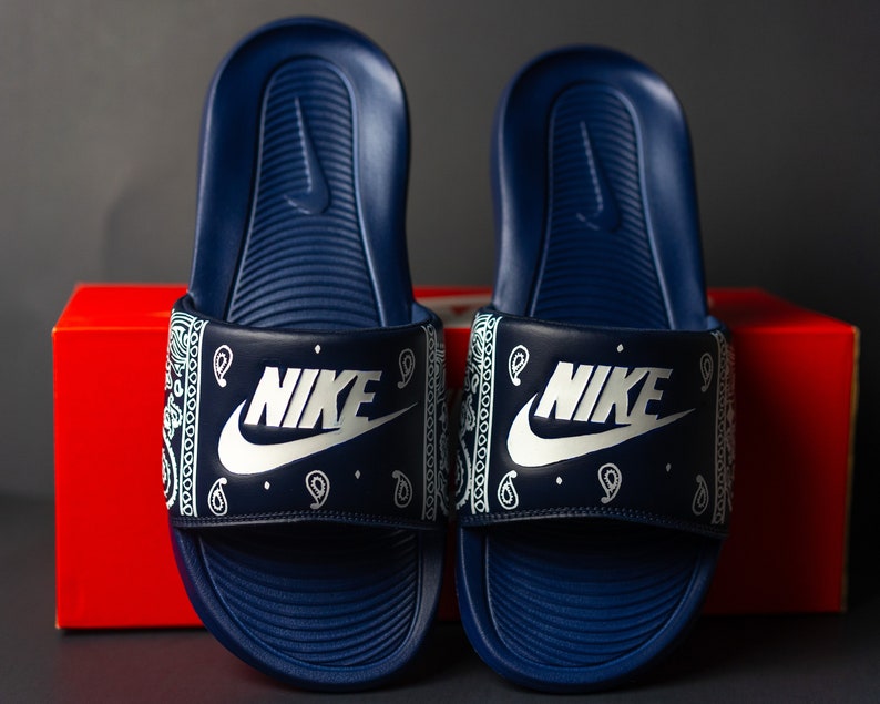 May include: A pair of navy blue Nike slides. The slides feature a white paisley pattern on the strap with the Nike logo in white. The slides are displayed in front of a red box.