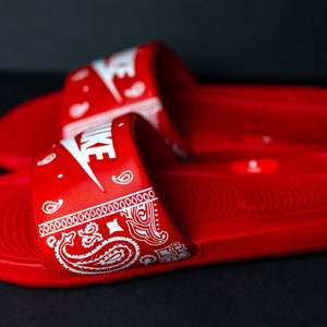 May include: Pair of red Nike slides with white Nike logo and a white paisley design on the strap. The slides are made of a smooth material and are designed for comfort and style.