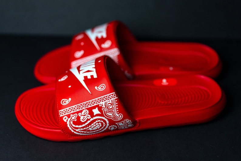 May include: Pair of red Nike slides with white Nike logo and a white paisley design on the strap. The slides are made of a smooth material and are designed for comfort and style.