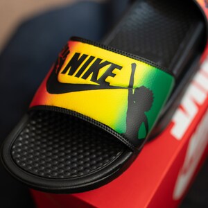 K&ouml;nnte beinhalten: Schwarze Nike-Badesandalen mit gelbem, gr&uuml;nem und rotem Design. Das Nike-Logo ist schwarz. Die Sandale zeigt eine Silhouette einer Person mit erhobenem Arm.