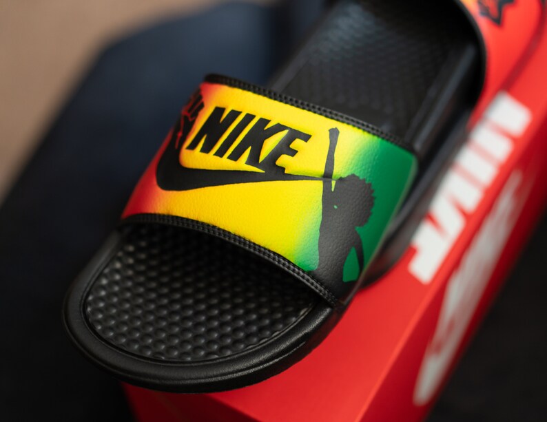 K&ouml;nnte beinhalten: Schwarze Nike-Badesandalen mit gelbem, gr&uuml;nem und rotem Design. Das Nike-Logo ist schwarz. Die Sandale zeigt eine Silhouette einer Person mit erhobenem Arm.