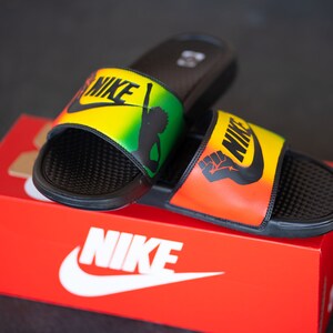 K&ouml;nnte beinhalten: Schwarze Nike-Slides mit gelbem, gr&uuml;nem und rotem Design. Die Slides zeigen das Nike-Logo und eine Grafik eines erhobenen F&auml;ustlings. Die Slides liegen auf einer roten Nike-Box mit dem Nike-Logo.