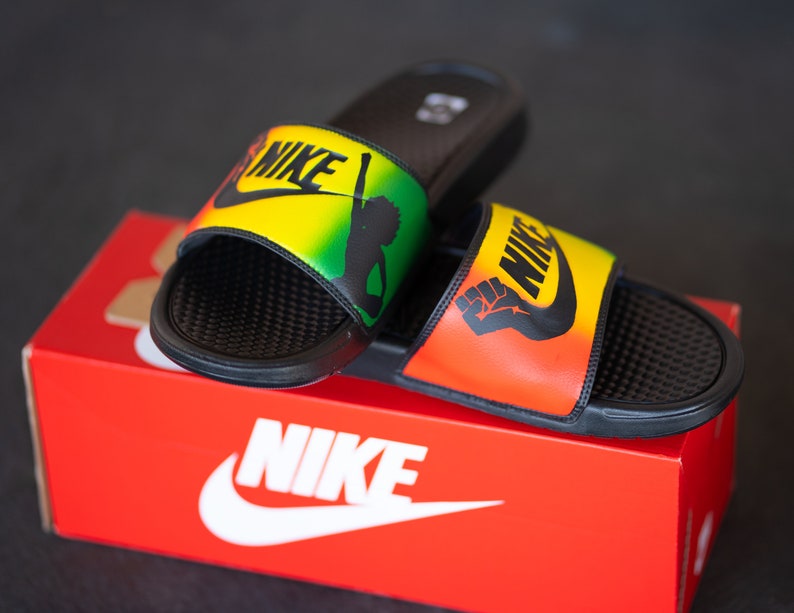 K&ouml;nnte beinhalten: Schwarze Nike-Slides mit gelbem, gr&uuml;nem und rotem Design. Die Slides zeigen das Nike-Logo und eine Grafik eines erhobenen F&auml;ustlings. Die Slides liegen auf einer roten Nike-Box mit dem Nike-Logo.