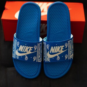 May include: Pair of blue Nike slides with white paisley pattern straps and the Nike logo. The slides are on a black surface with a red Nike box in the background.